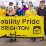 Photo of Disability Pride in Brighton, with people holding up a sign, including Caroline Lucas MP and the mother who began the movement